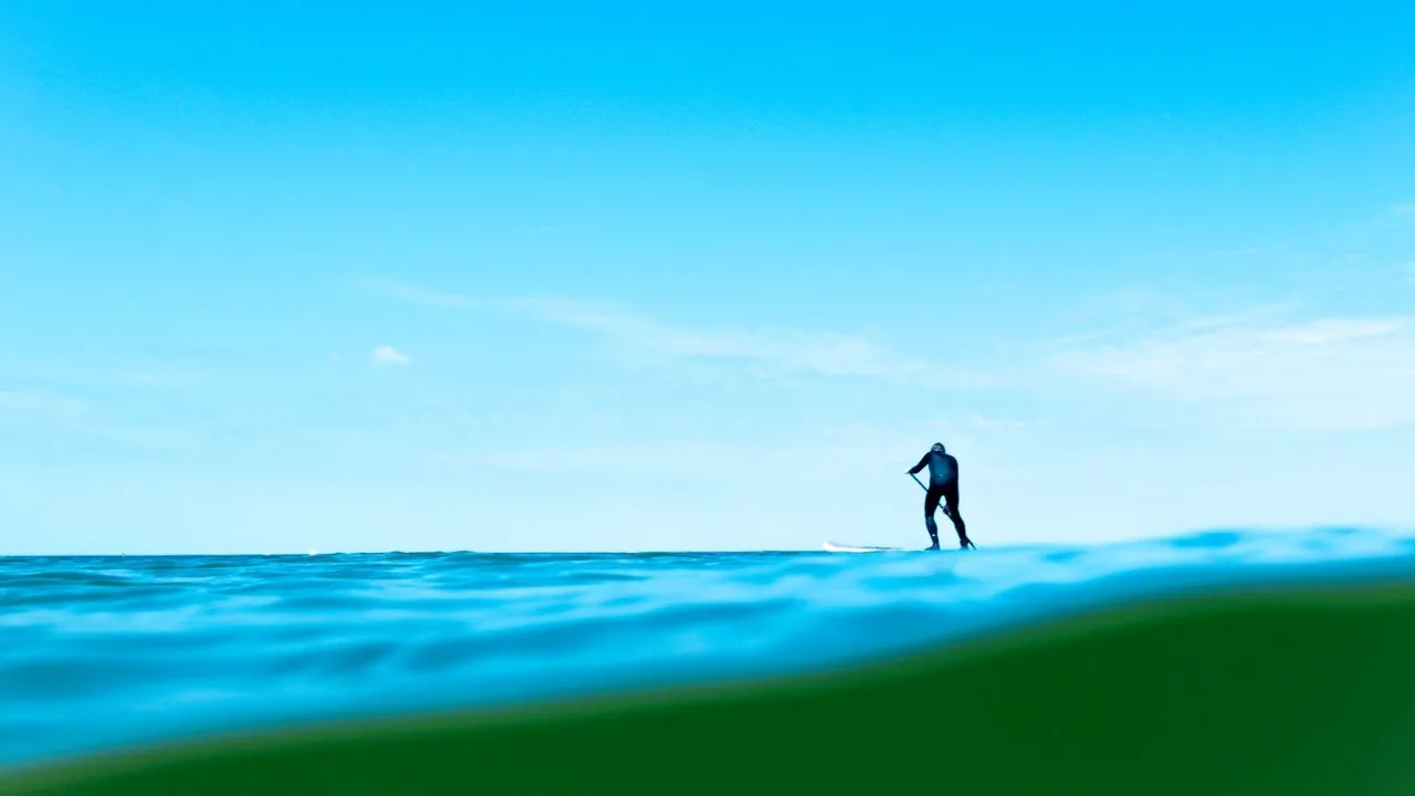 A paddleboarder out on open water against a bright sky, mid-stroke and moving.