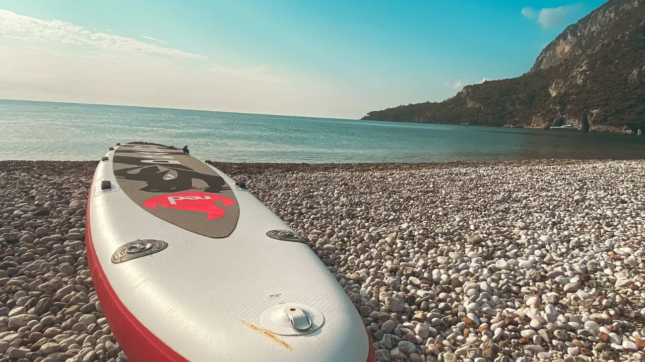 An inflatable paddleboard resting on a pebble beach, a calm bay and headland behind.
