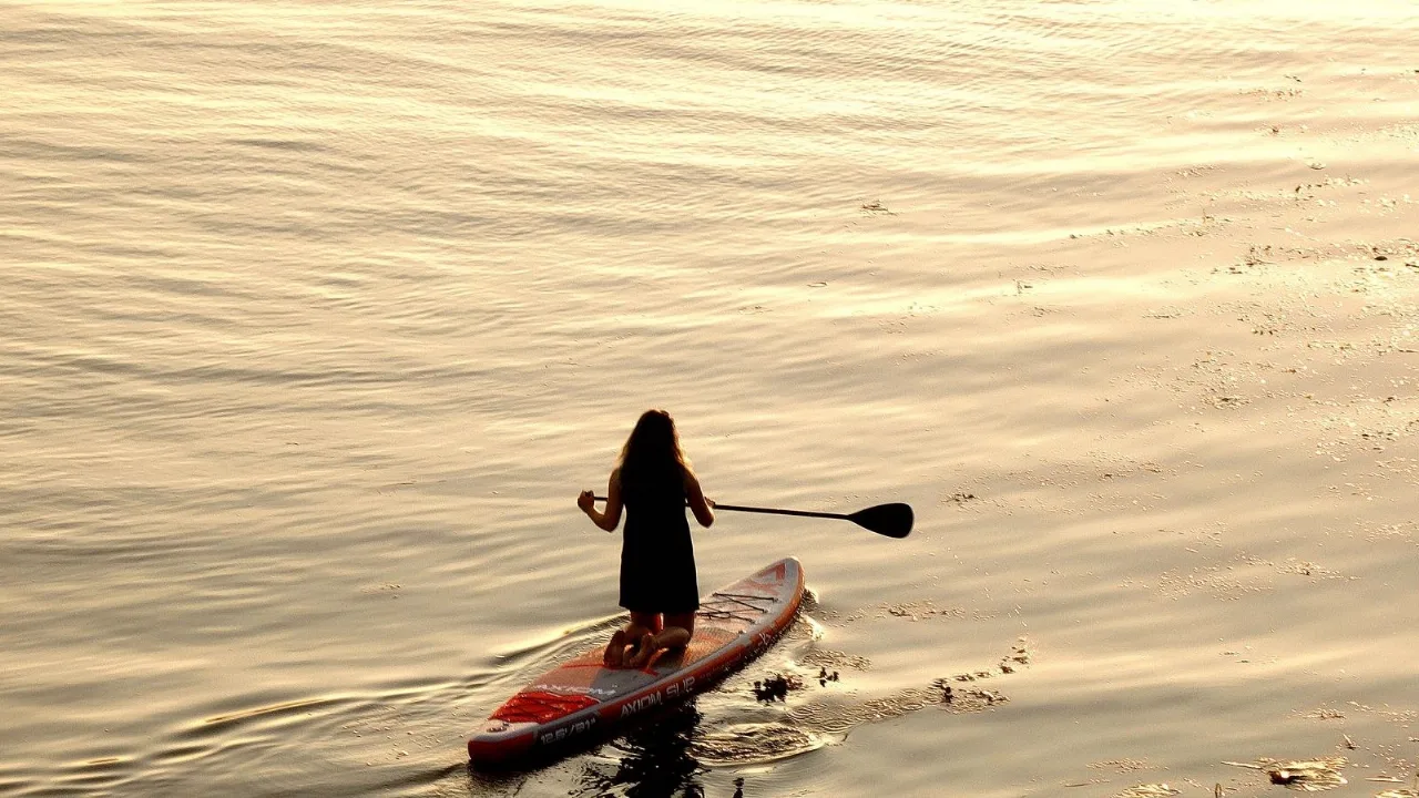 A paddler kneeling on their board on calm, warm-lit water at golden hour, drifting quietly.