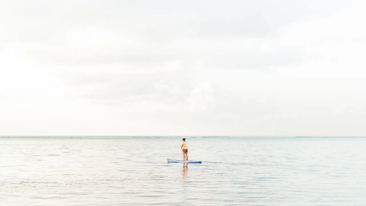 A lone paddleboarder standing on their board far out on calm, pale open water under a soft overcast sky.